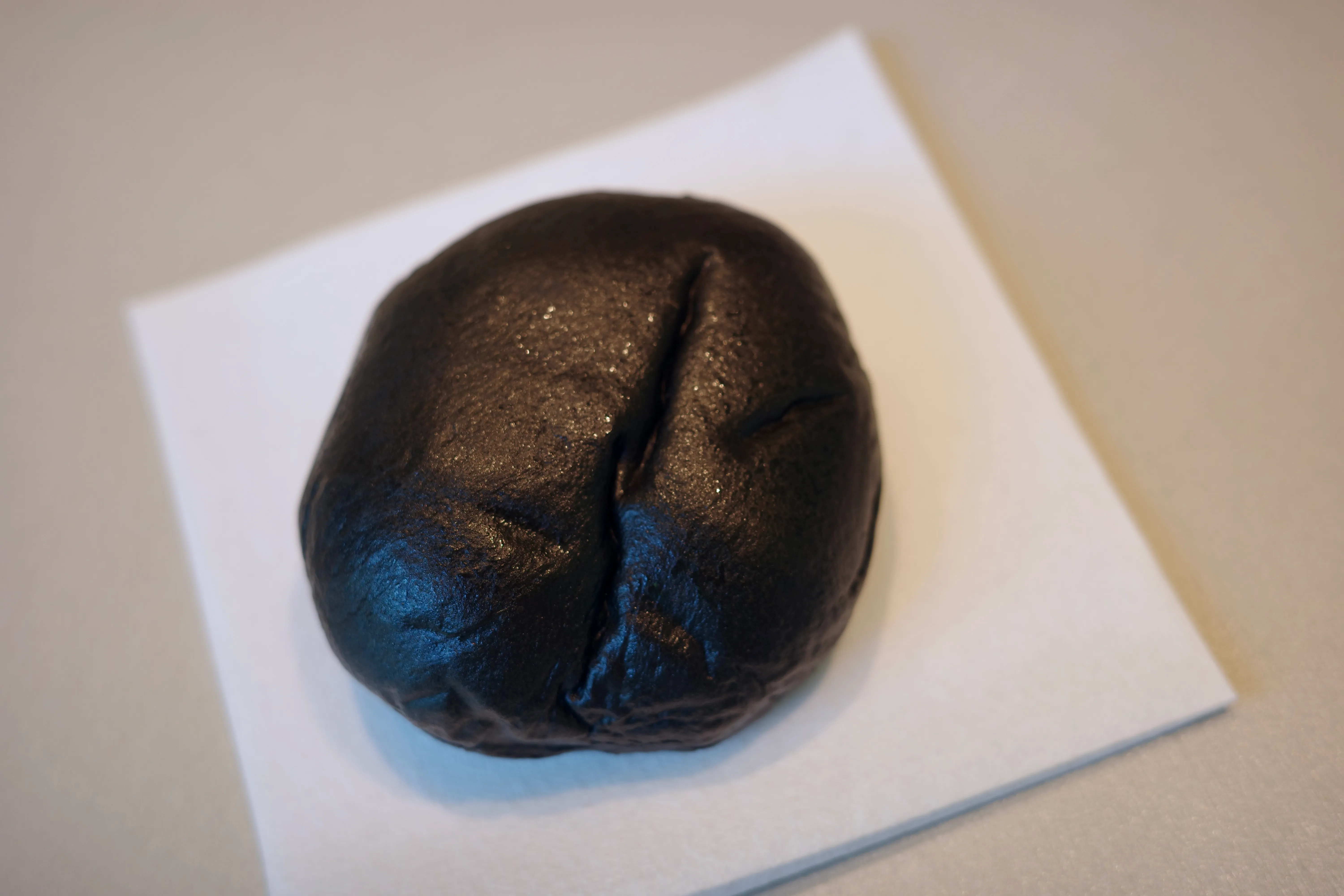 A whole charcoal-black mochi bun on white parchment, glossy and round, before the first bite