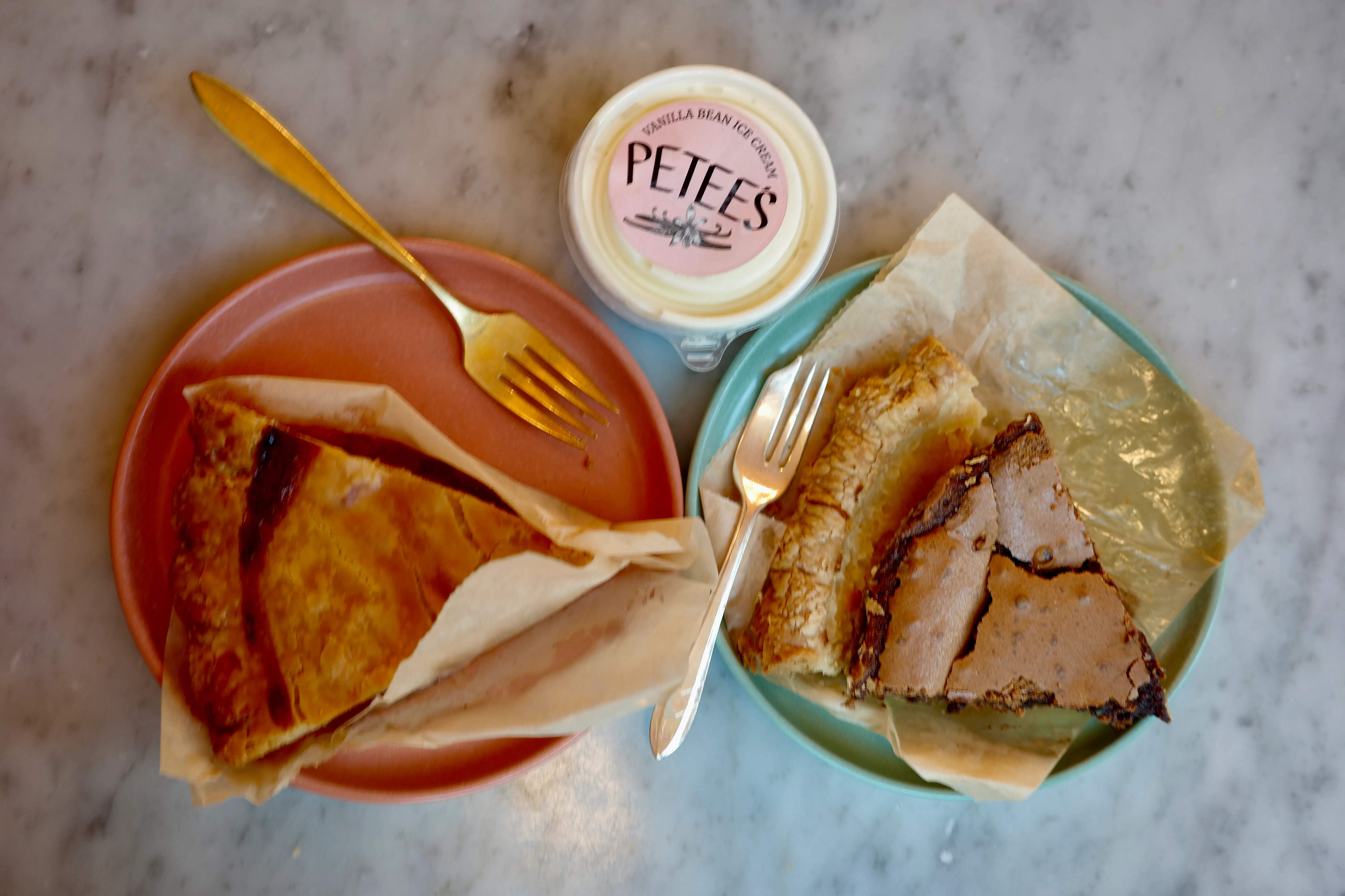 Overhead spread on marble: a folded pie slice on a coral plate, the chocolate chess on a teal plate, and a cup of Petee's vanilla bean ice cream between them