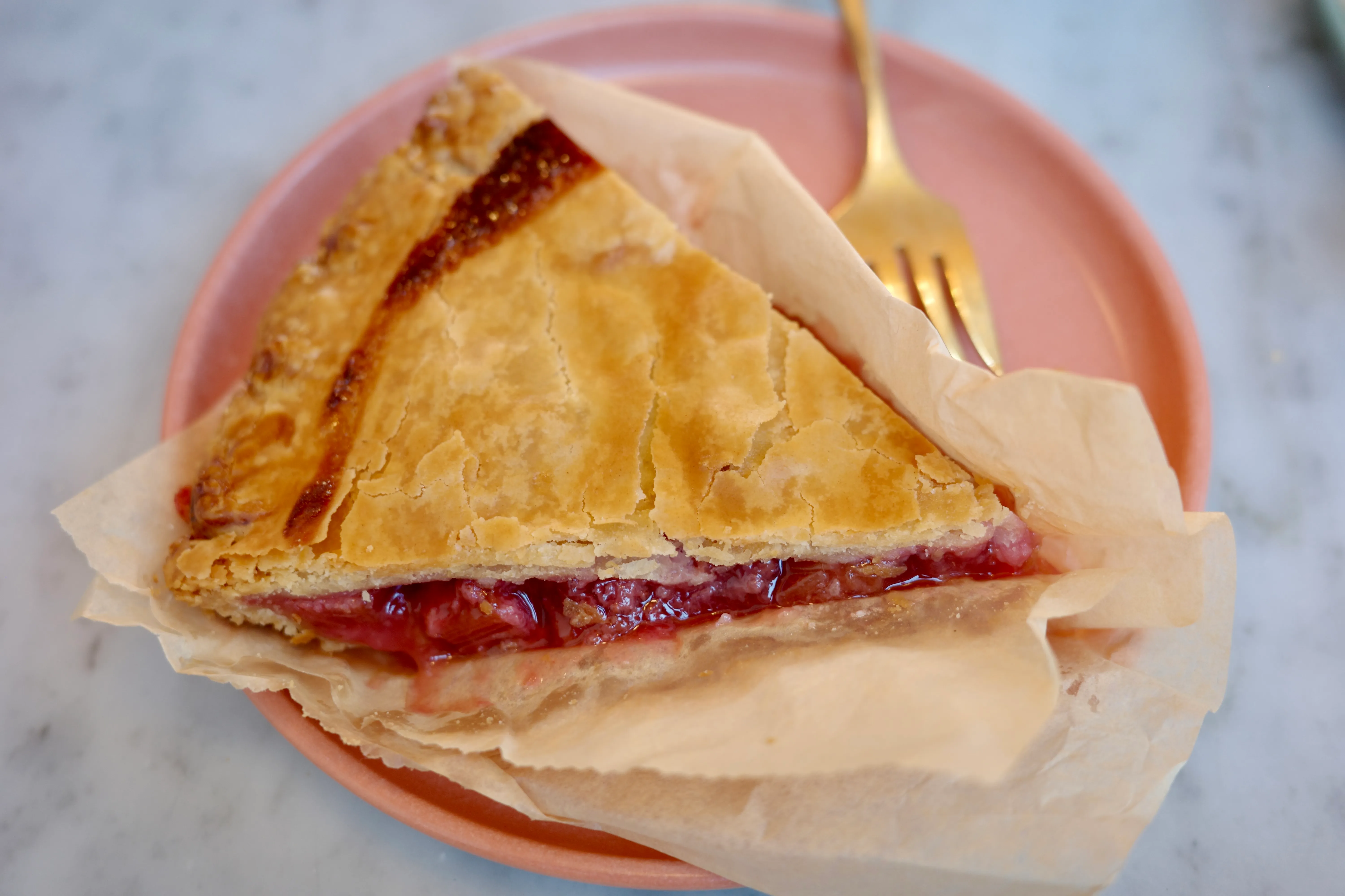 A slice of strawberry rhubarb pie on a pink plate, flaky golden crust with bright red filling, gold fork alongside